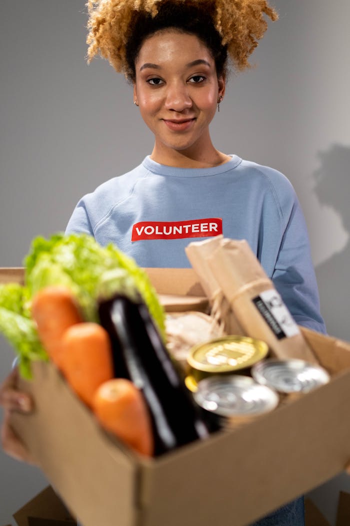 Smiling woman holding a box filled with various vegetables and canned goods during a volunteer drive.
